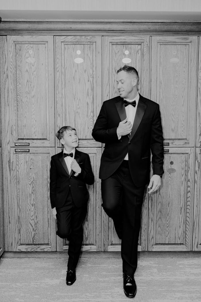 gallery-02 Black and white photo of a father and son in matching tuxedos, indoors.